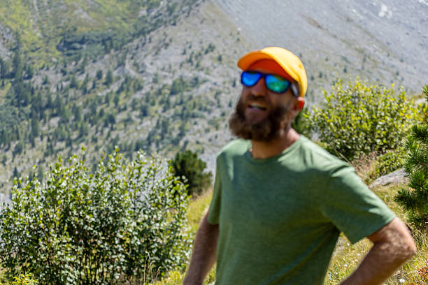Bearded man in green shirt and orange cap hiking outdoors in mountainous terrain. Active adult enjoying nature on a sunny day. Outdoor adventure and fitness lifestyle concept.