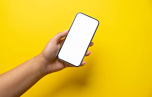Close-up of a female hand holding a modern smartphone with a blank white screen against a solid yellow background