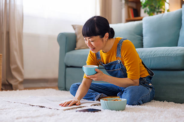 Beautiful Asian woman sitting on the living room floor, reading newspaper, drinking coffee and eating cookies while enjoying leisure time and relaxing at home