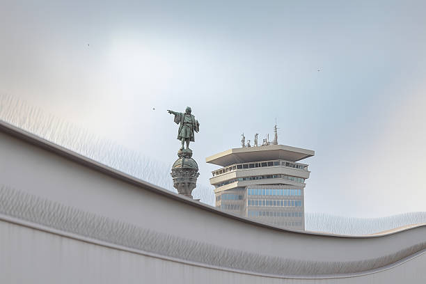 View of the Christopher Columbus Monument with the World Trade Center building in the background, seen from the waterfront in Barcelona, Spain. The image captures the iconic statue pointing towards the sea, framed by modern architecture under a soft sky.