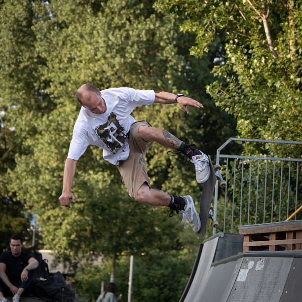 Constance, Germany – 7.9.2022: A man executes a skateboard trick on an outdoor ramp. He's wearing a white t-shirt and beige shorts, with sneakers, in mid-air, demonstrating dynamic movement. The ramp is metal and wood, and there are trees surrounding the area providing a natural backdrop. A seated onlooker is visible in the background, observing the performance. The scene reflects an active, outdoor skateboarding environment.