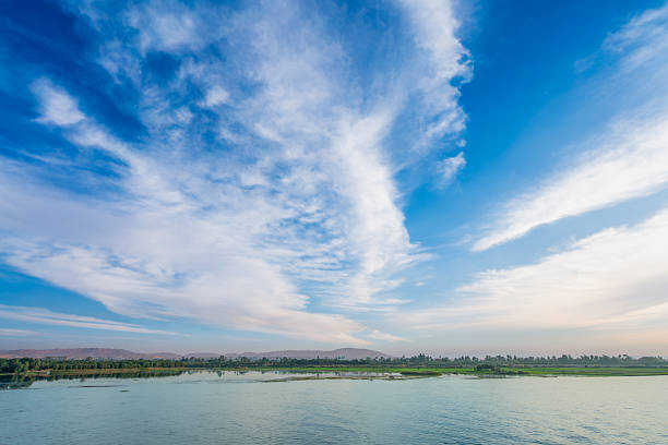 A dramatic sky with long, wispy white clouds over the calm waters of the Nile River. This serene landscape view was captured during a cruise after passing the Esna Lock.
