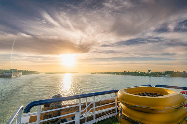 A serene view of the Nile River at sunset, seen from a cruise ship deck. The golden light reflects on the water, with life rafts and guardrails visible in the foreground.