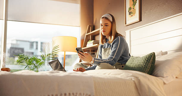 istock Woman, student and phone with laptop on bed, texting and check notification with web chat in home. Girl, smartphone and computer for elearning, study or virtual course for college assessment in house 2230317464
