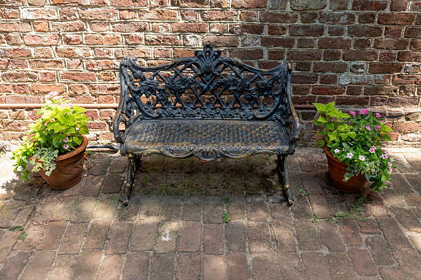 Charleston, South Carolina, USA - July 1, 2025: Decorative cast iron bench flanked by potted flowers against historic brick wall at Middleton Place Plantation.