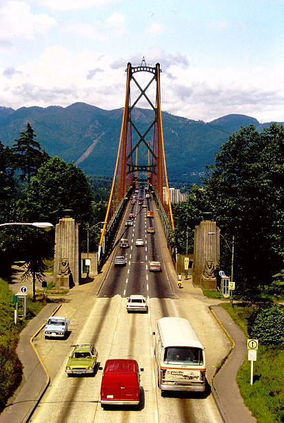 Suspension bridge with orange cables and green steel structures, vehicles in motion, and mountain backdrop in Vancouver, British Columbia. Scanned from 1977 slide film.