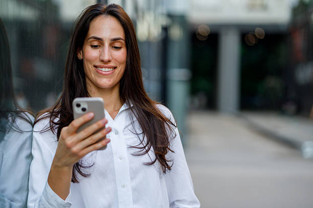 istock Young businesswoman smiling while using a smartphone in front of a sleek, modern office building, radiating confidence and happiness 2230343039