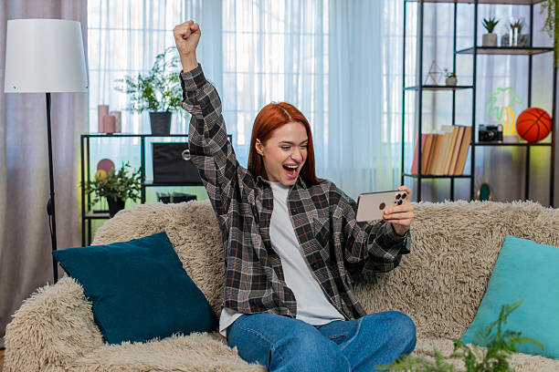 istock Woman sits on sofa playing mobile game excitedly tapping screen celebrating fun victory and success 2230347435