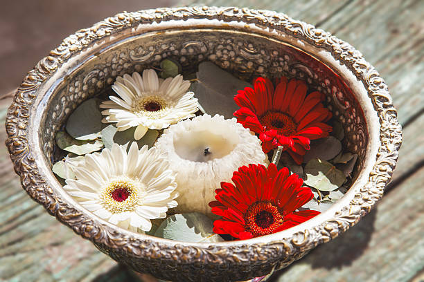 Antique silver bowl is filled with water, eucalyptus leaves, a floating white candle, and red and white daisies, creating a serene and decorative centerpiece