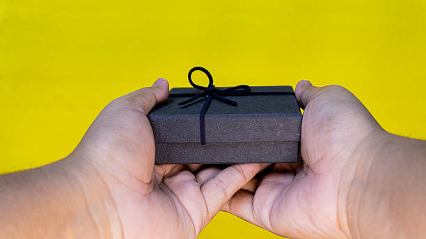 istock Closed black gift box offered with both hands on a yellow backdrop, ideal for gifting, luxury, and festive celebration concepts. 2230368714