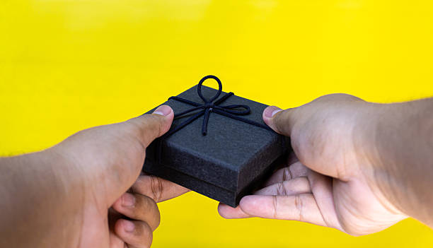 istock Closed black gift box offered with both hands on a yellow backdrop, ideal for gifting, luxury, and festive celebration concepts. 2230368722