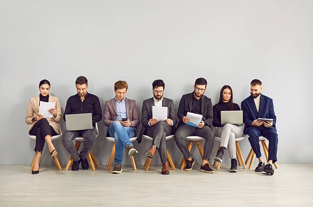 Happy young professional business people, group of applicants are sitting on chairs, waiting for interview, sitting in line with laptops, notebooks and other resources. stock photo