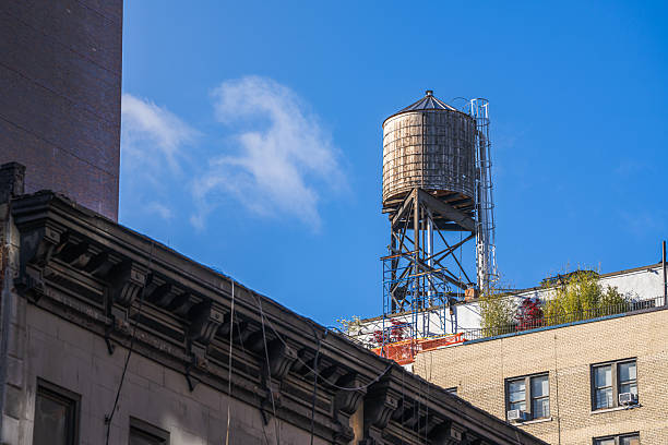 An old metal water tower rises above a patchwork of faded brick and modern facades, anchored by a stark black clock on a minimalist beige wall.