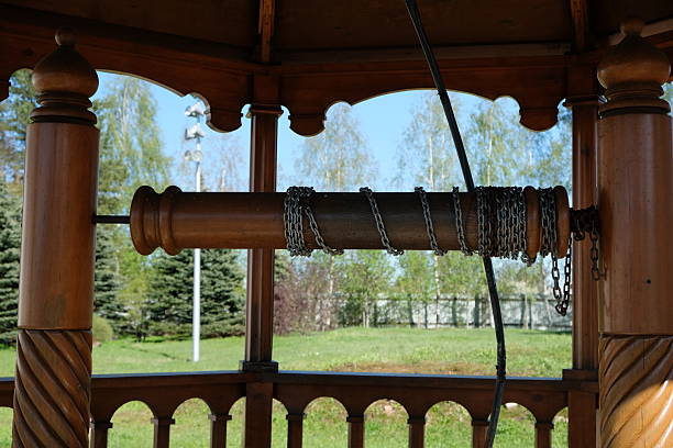 A wooden gazebo features a decorative central post surrounded by chains, set against a vibrant green park background under a clear blue sky during daylight.