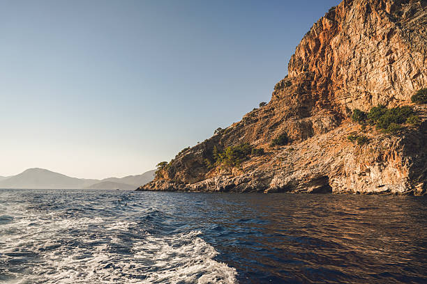 Scenic coastal view with rocky cliffs and trees, captured from a boat moving across the blue sea. Mountain silhouettes appear in the background under a clear blue sky. A serene scene reflecting summer, nature, and tranquility.