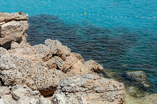 Coastal rocks with vibrant turquoise sea. View of the Mediterranean coastline, with a clear demarcation between the shallow and deep water. Coastal rocks jutting out into the clear sea