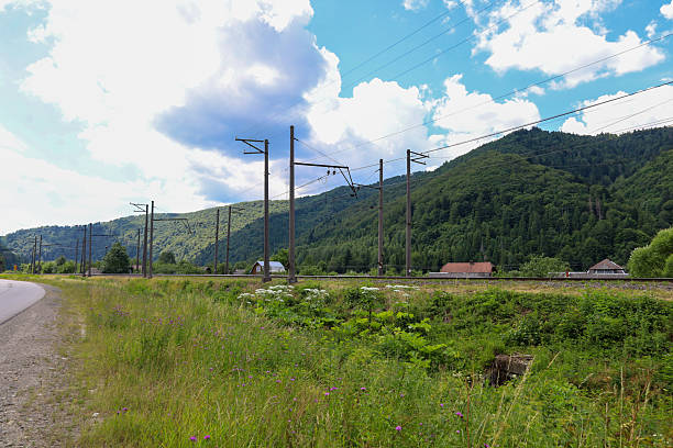 istock Railway tracks and power lines run along hills covered with a dense forest. An automobile road runs nearby, and against the background of the blue sky with white clouds, small houses are visible, creating a picture of rural life. 2230406020