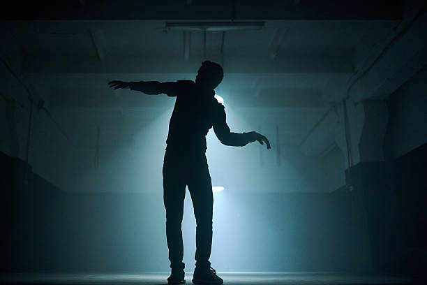 Handsome bearded young man in black clothing (jeans, shirt and sport shoes) is dancing modern street dance, backlit, silhouette of a dancer. Shooting in big room with cone-shaped lamps on a black background