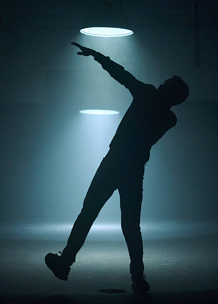 Handsome bearded young man in black clothing (jeans, shirt and sport shoes) is dancing modern street dance, backlit, silhouette of a dancer. Shooting in big room with cone-shaped lamps on a black background