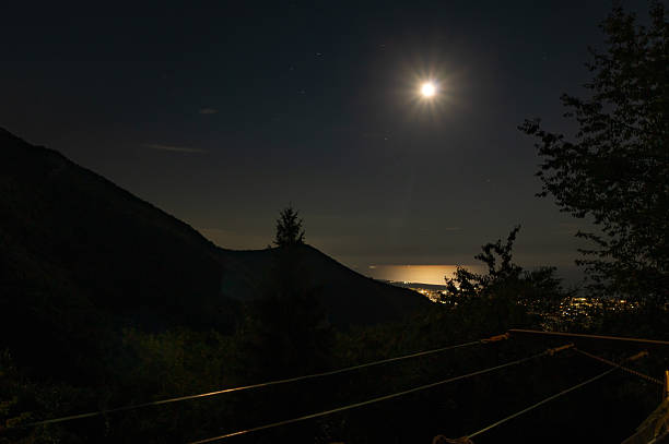 Night view of bright moon shining over sea and illuminated coastal cityscape, seen from mountain viewpoint.