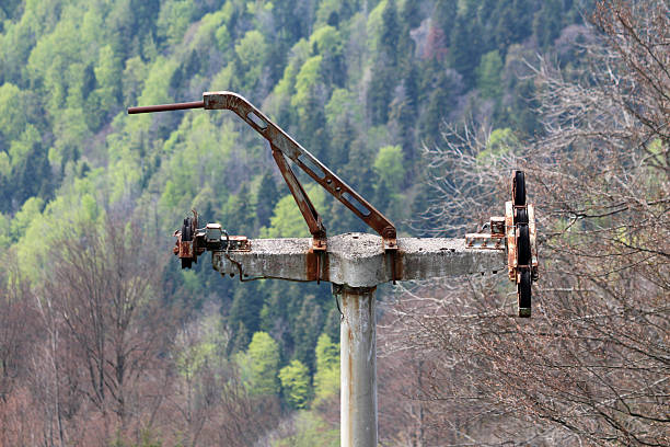 Old ski lift support made of concrete and rusty metal with pulley wheels stands on mountain slope, framed by bare winter trees and dense green forest backdrop
