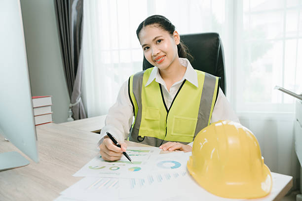 istock Female construction worker in safety gear reviewing project documents at desk with computer and safety helmet nearby 2230423054