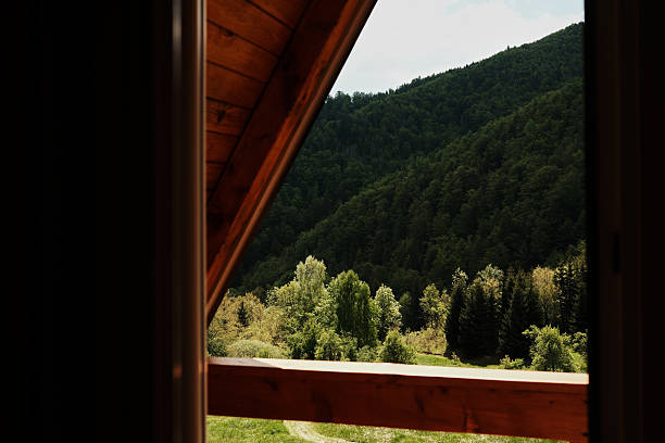 Looking through a triangular attic window toward green wooded hills. A-frame cabin in Tara National Park, Serbia. Peaceful nature and architectural charm combined.