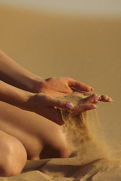 Close-up view of hands gracefully releasing fine sand amidst a serene desert backdrop. The warm tones of the scene evoke tranquility and natural beauty, depicting a moment of mindfulness and connection with nature. Perfect to illustrate concepts of time, impermanence, and desert landscapes.