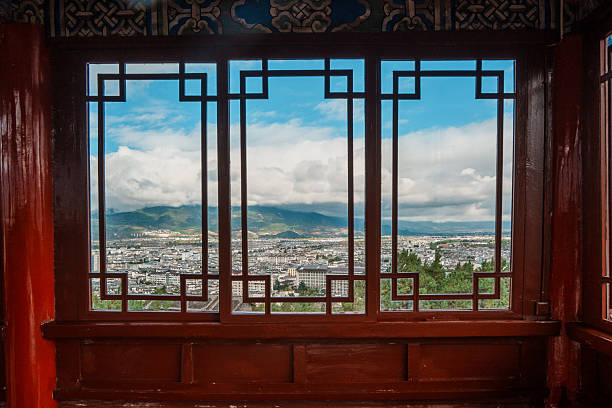 Scenic view of Lijiang city and surrounding mountains framed through a traditional Chinese window in Wangu Pavilion, Yunnan.