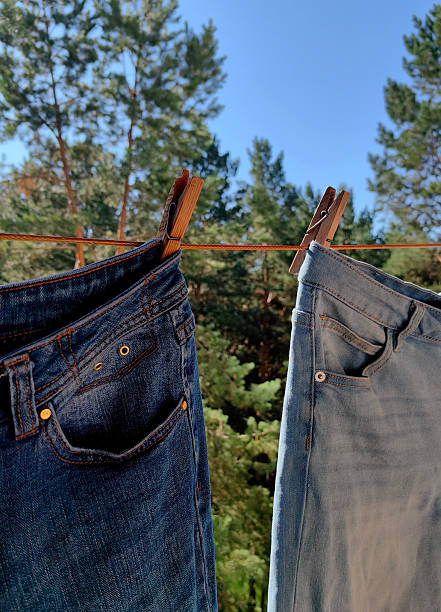 two pairs jeans are hanging on clothesline, swaying gently in breeze. tall green trees and bright blue sky form serene outdoor backdrop, creating calm atmosphere. close up.