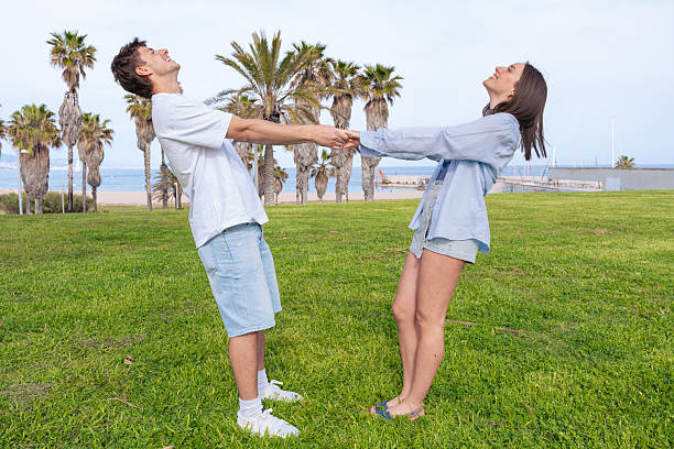 Joyful young couple standing on green grass in a park near the beach, holding hands and leaning backward while smiling, enjoying a playful and fun outdoor moment together with palm trees