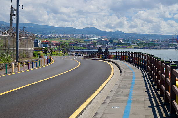 A rainbow-painted fence lines the HaguAewol Coastal Road as it descends from Eoyeong Park toward Sasu Port, with a blue bike lane tracing the seawall above basalt rocks.