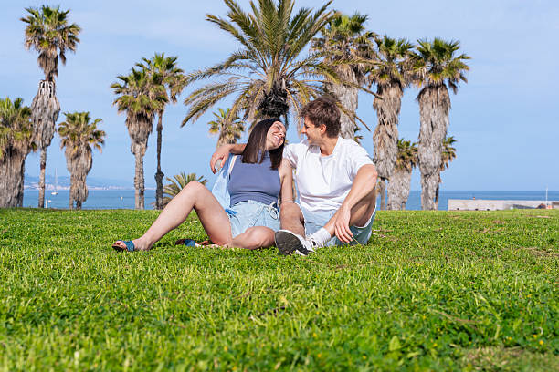 Young couple sitting on green grass under palm trees at the beach, enjoying a sunny summer day, smiling and looking at each other with affection and happiness, romantic outdoor scene by the ocean