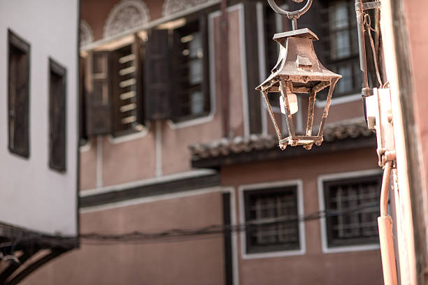 Historic metal lantern mounted on a building wall in Plovdiv Old Town, Bulgaria, with background view of traditional Bulgarian Revival houses featuring wooden shutters and decorative facades.