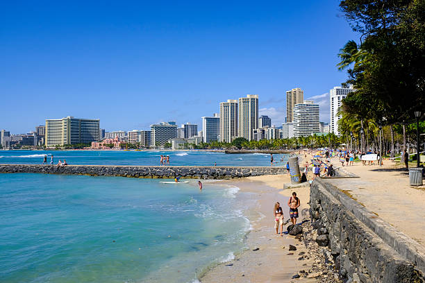 Waikiki, Hawaii, February 18, 2024:  Sunlit waves meet the bustling charm of Waikiki, where beachgoers stroll along the shore under a backdrop of palm trees and high-rise hotels.