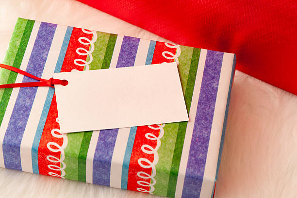 Small wrapped striped Christmas present with blank white gift tag on white fur background. Cropped metallic red wrapped box in background.