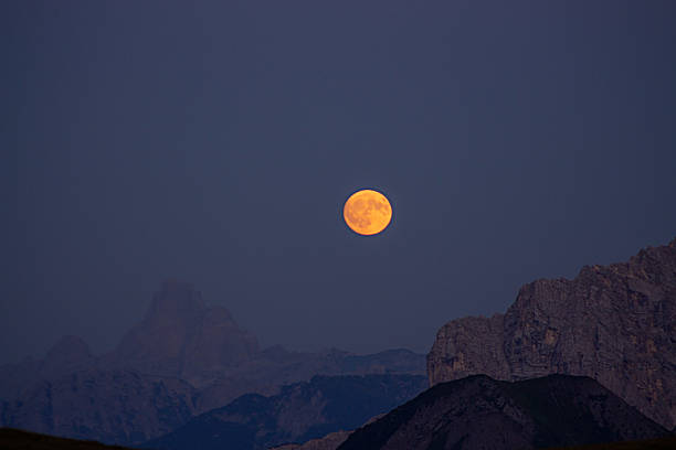 A dramatic and captivating close-up shot of a luminous full moon glowing over the Dolomites, captured from the Mondeval plateau at twilight. The photograph features the large, golden-orange moon as a stunning focal point in a dark blue-gray sky. Below and around the moon, the rugged peaks of the Dolomites are in a powerful silhouette, creating a beautiful and mystical contrast. This image perfectly conveys the awe-inspiring and magical beauty of the alpine landscape at night, making it ideal for projects related to travel, nature, landscape photography, and themes of astronomy and serenity.