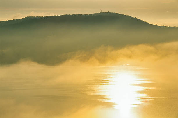 The golden light of sunset falling through dark clouds onto the water, turning it golden. A forest-covered hillside is in silhouette against the dark sky.