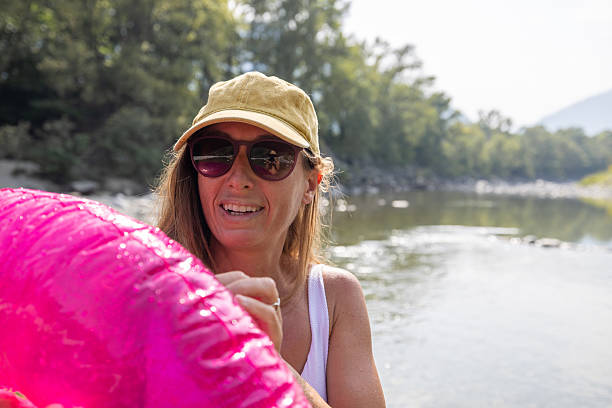 Smiling woman wearing sunglasses and cap holds a bright pink inflatable pool float by the river. Outdoor summer fun with water recreation and leisure activities. Relaxing nature scene.