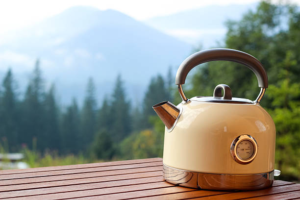 A stylish kettle rests on a wooden table set against a backdrop of mountains and evergreen trees during the morning.