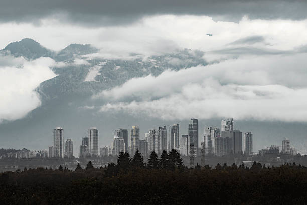 Urban skyline captured under dramatic weather, with clouds surrounding tall buildings and distant mountains, creating a scene of nature blending with modern architecture and emphasizing the contrast between landscapes and structures.