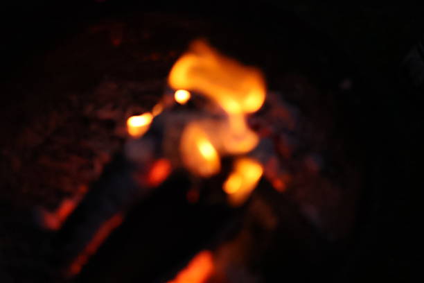 A close-up, low-light photograph capturing the intimate and warm glow of a campfire. The image focuses on a stack of burning wood logs, with bright, dancing yellow and orange flames licking at the textured bark.