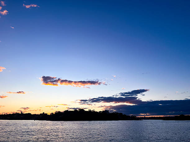 Over a city harbour, a towering nimbus cloud formation glows with pink and salmon tones, dramatically illuminated by the fading light of a beautiful sunset