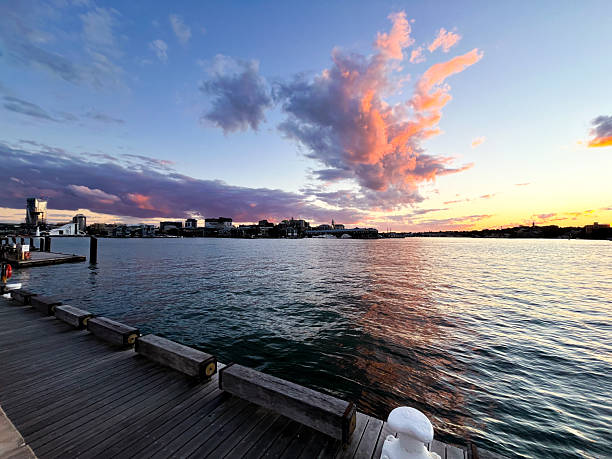 Over a city harbour, a towering nimbus cloud formation glows with pink and salmon tones, dramatically illuminated by the fading light of a beautiful sunset