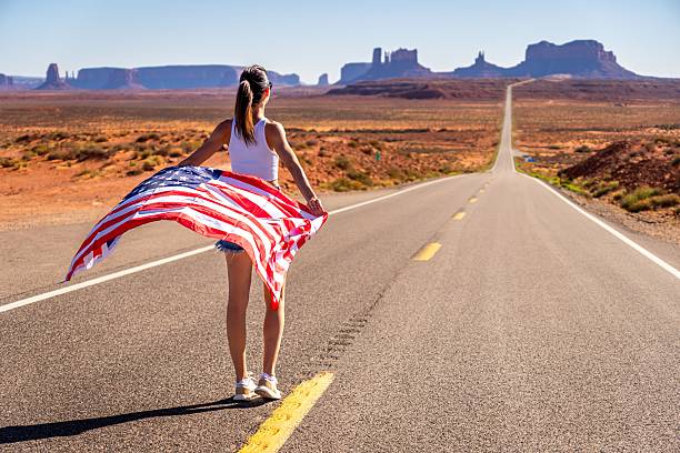 Young woman holding American flag on a desert road in Monument Valley, Utah, showcasing freedom and adventure.
