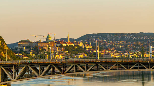 Panoramic view of Budapest's Chain Bridge crossing the tranquil Danube River, with the majestic Buda Castle and Royal Palace illuminated by warm sunset light against rolling hills.