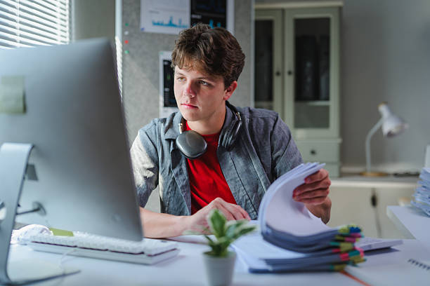 istock Teenage student studying intently, typing on laptop while organizing documents in minimalist home workspace 2230538515