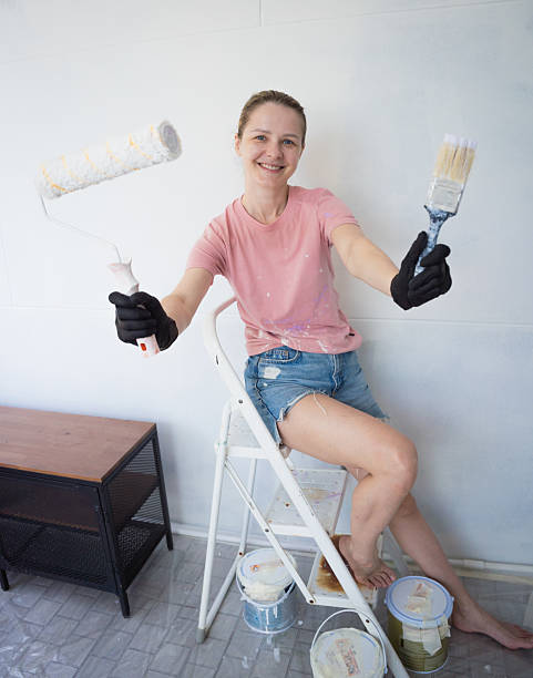 istock Young woman painting indoors wall with roller using stepladder 2230542490