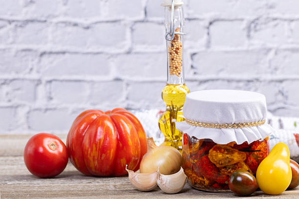Glass jar with sun dried red, black and yellow cherry tomatoes in olive oil and with herbs on the rustic brown wooden table. A bottle with oil and herbs, garlic and various tomatoes on the background.