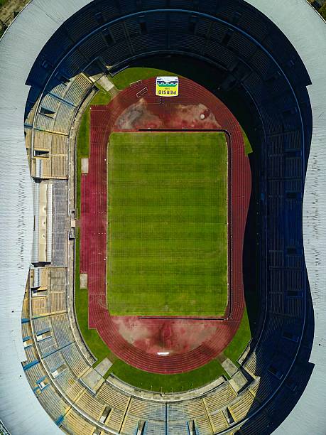 This aerial photograph captures Gelora Bandung Lautan Api (GBLA) Stadium, the home ground of Persib Bandung, located in Bandung, West Java, Indonesia. The massive football stadium, with its modern oval design and large seating capacity, stands prominently amidst the urban and green landscapes surrounding it. The lush green football pitch and running track are clearly visible at the center, while the grand stands encircle the field in a striking architectural form. Beyond the stadium, housing areas, rice fields, and the iconic Al Jabbar Grand Mosque can also be seen, reflecting the blend of sports, culture, and community in Bandung. As one of the largest stadiums in Indonesia, GBLA is not only a landmark for football fans but also a symbol of pride for the city of Bandung and its people